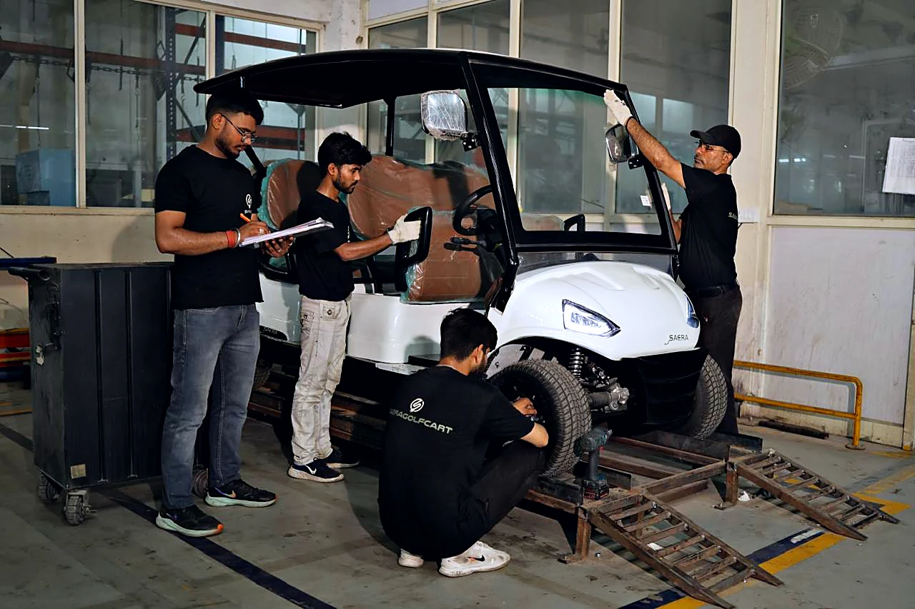 Saera technicians assembling a white electric golf cart on the factory floor — one fitting the front wheel, one checking a build sheet, one aligning the windscreen.